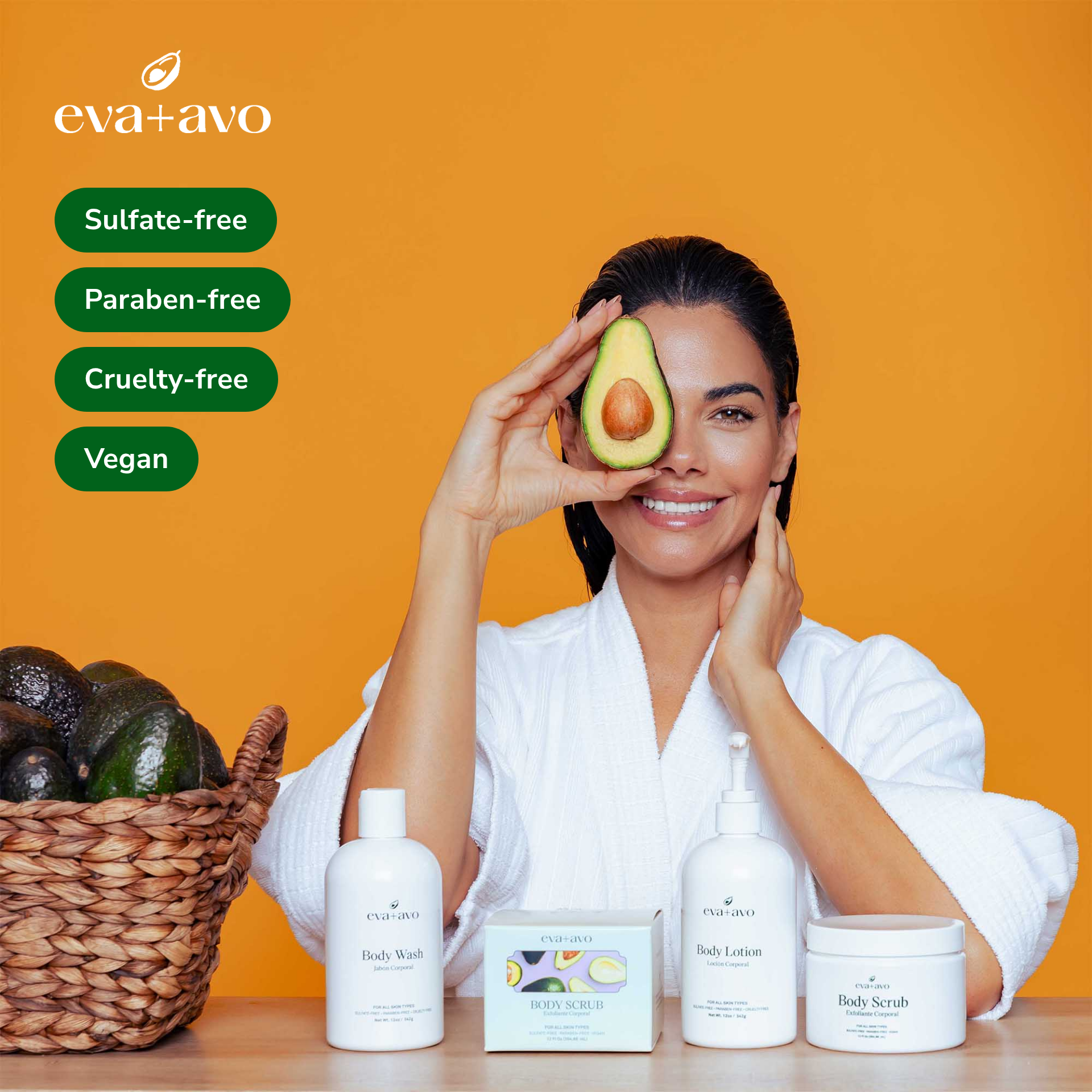 Woman in a white robe holds half an avocado over one eye against an orange background, beside four green labels reading Sulfate-free, Paraben-free, Cruelty-free and Vegan, with Eva+Avo body wash, scrub, lotion and a basket of avocados in front of her.